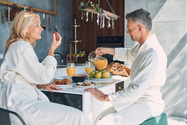 国内のキッチンで時間を過ごしながら一緒に朝食を楽しむバスローブの美しい成熟したカップル — ストック写真