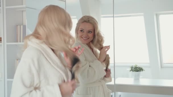 Mujer joven en albornoz peinando el cabello mientras está de pie frente al espejo — Vídeos de Stock