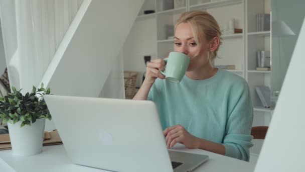 Hermosa mujer joven bebiendo café mientras usa el ordenador portátil en casa — Vídeo de stock