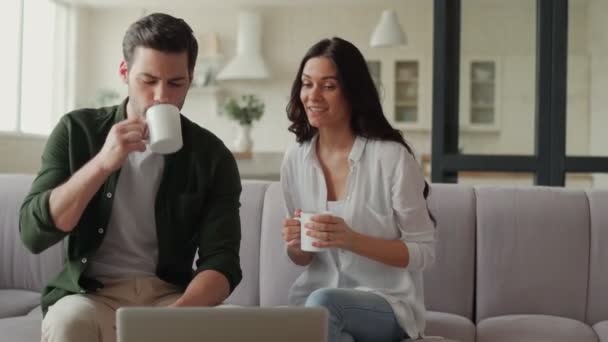 Joven pareja sonriente bebiendo café y utilizando el ordenador portátil mientras está sentado en el sofá — Vídeos de Stock