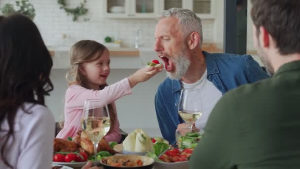 Klein meisje voeden haar grootvader en glimlachen tijdens het eten met familie — Stockvideo