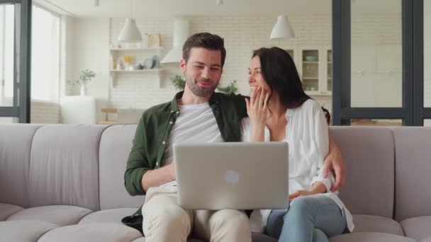 Junges lächelndes Paar trinkt Kaffee und benutzt Laptop, während es auf dem Sofa sitzt — Stockvideo