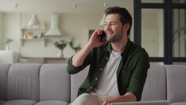 Красивый молодой человек разговаривает по смартфону и улыбается, сидя на диване — стоковое видео