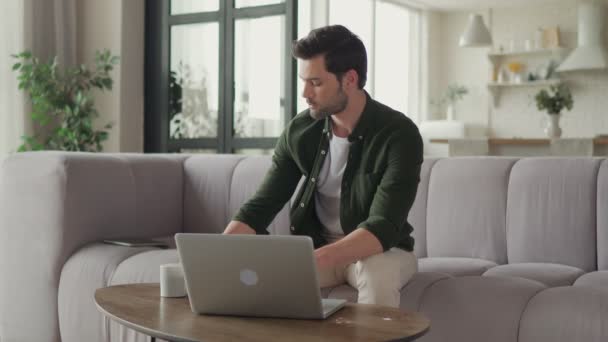 Молодой человек пользуется ноутбуком и пьет кофе, сидя дома на диване — стоковое видео