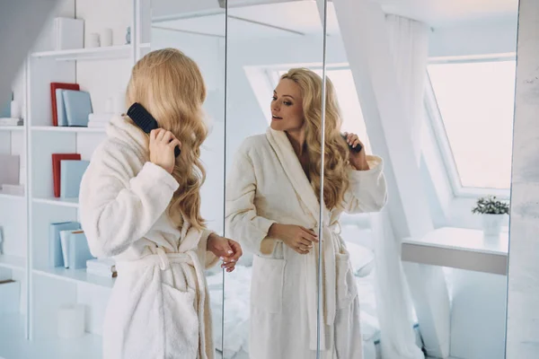 Приваблива молода усміхнена жінка в халаті розчісує волосся, стоячи перед дзеркалом вдома — стокове фото