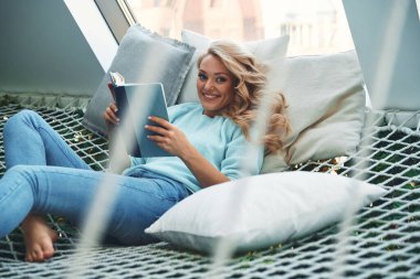 Güzel genç bir kadın kitap okuyor ve gülümsüyor evde büyük bir hamakta dinlenirken.