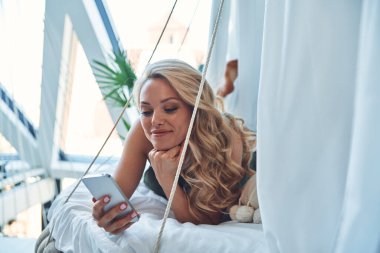 Güzel genç bir kadın akıllı telefon kullanıyor ve evde yatakta dinlenirken gülümsüyor.