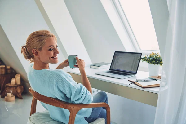 Achteraanzicht van jonge glimlachende vrouw genieten van warme drank tijdens het werken thuis — Stockfoto