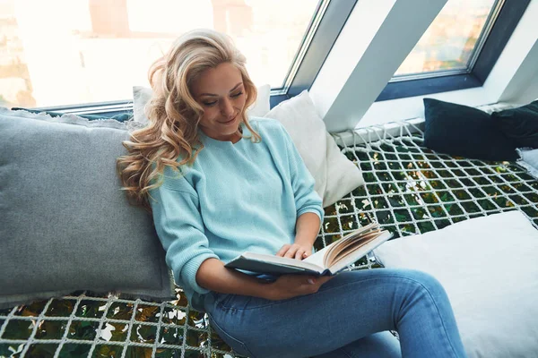 Mooie jonge vrouw lezen boek andd glimlachen terwijl ontspannen in grote hangmat thuis — Stockfoto