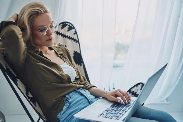 Attraktiv ung kvinna använder laptop medan du sitter i bekväm stol hemma — Stockfoto