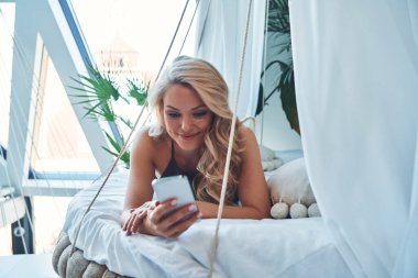 Güzel genç bir kadın akıllı telefon kullanıyor ve evde yatakta dinlenirken gülümsüyor.