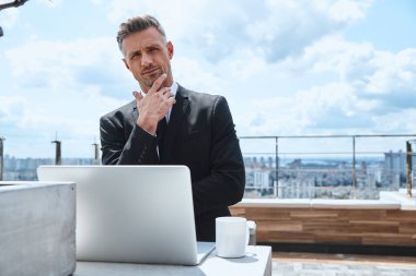 Kendine güvenen olgun bir adam dizüstü bilgisayarda çalışıyor ve çatıda kameraya bakıyor.