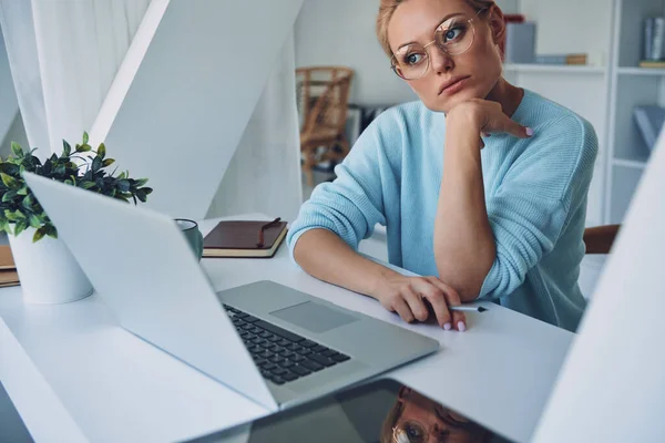 Belle jeune femme utilisant un ordinateur portable et regardant réfléchi tout en travaillant au bureau — Photo