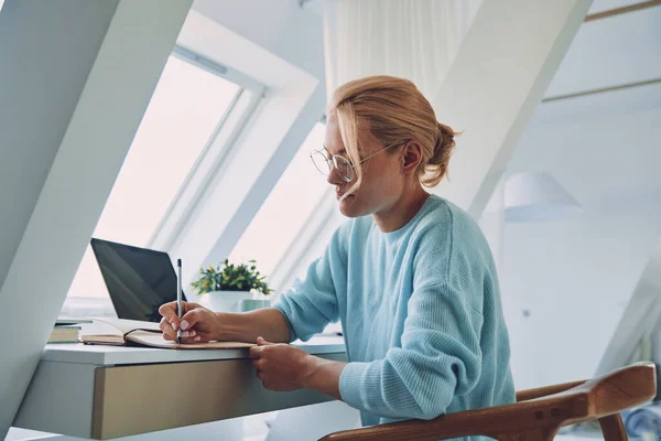 Gyönyörű fiatal nő ír valamit jegyzetfüzetbe, miközben az irodában dolgozik. — Stock Fotó