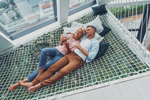 Beautiful mature couple examining smart phone and smiling while relaxing in big hammock at home together — Stock Photo, Image