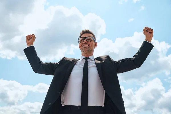 Heureux homme mature en costume complet gardant les bras levés tout en se tenant devant le ciel bleu à l'extérieur — Photo