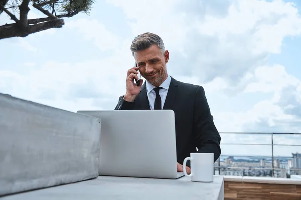 Hombre maduro de confianza trabajando en el ordenador portátil y hablando en el teléfono móvil mientras está de pie en la terraza de la azotea — Foto de Stock