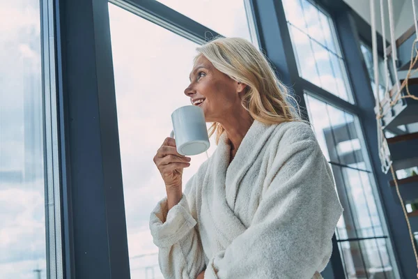 Schöne reife Frau in Bademänteln, Kaffee trinkend und lächelnd, während sie zu Hause am Fenster steht — Stockfoto