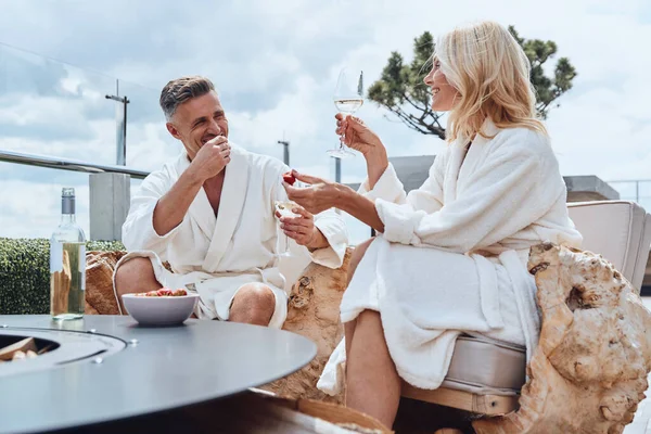Beautiful mature couple in bathrobes enjoying fruits and wine while relaxing in luxury hotel outdoors — Stock Photo, Image