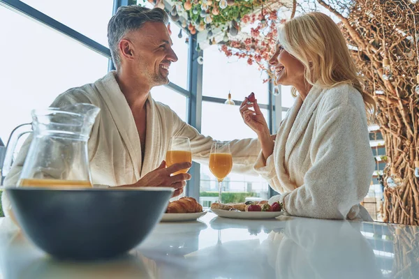 Vackra mogna par i badrockar njuter av frukost tillsammans medan de tillbringar tid i det inhemska köket — Stockfoto