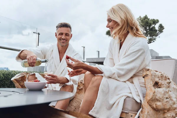Vackra mogna par i badrockar njuter av frukt och vin samtidigt som de kopplar av i lyxhotell utomhus — Stockfoto