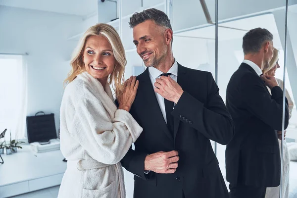 Mooie volwassen vrouw aanpassen pak van haar man terwijl zowel staan in de voorkant van de spiegel thuis — Stockfoto