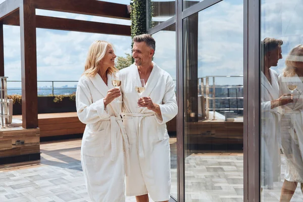 Beautiful mature couple in bathrobes enjoying champagne while relaxing in luxury hotel outdoors — Stock Photo, Image