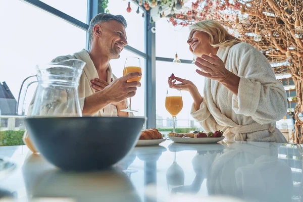 Vackra mogna par i badrockar njuter av frukost tillsammans medan de tillbringar tid i det inhemska köket — Stockfoto