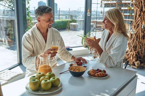 Vackra mogna par i badrockar njuter av frukost tillsammans medan de tillbringar tid i det inhemska köket — Stockfoto
