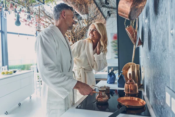 Vackra mogna par i badrockar tillagar kaffe tillsammans medan de tillbringar tid i köket — Stockfoto