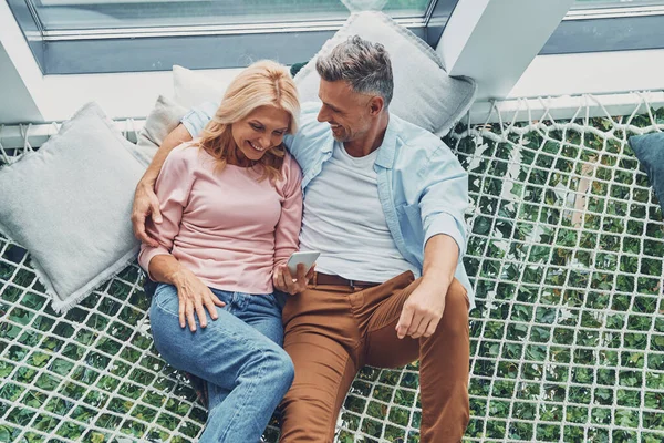 Vackra mogna par undersöker smart telefon och ler samtidigt som du kopplar av i stor hängmatta hemma tillsammans — Stockfoto