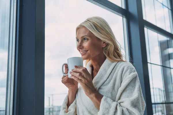 Красивая зрелая женщина в халатах пьет кофе и улыбается, стоя у окна дома — стоковое фото