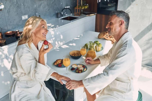 Top view of beautiful mature couple in bathrobes enjoying breakfast together while spending time in the domestic kitchen — Stock Photo, Image