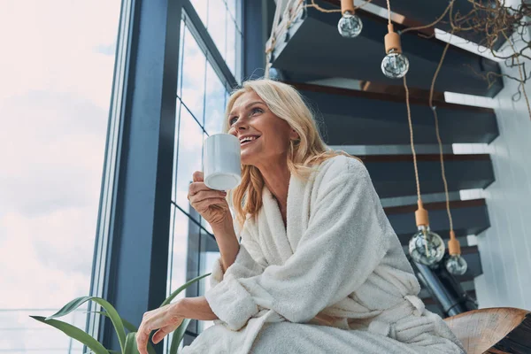 Повзрослевшая женщина в халате пьет кофе и улыбается, сидя у окна дома — стоковое фото