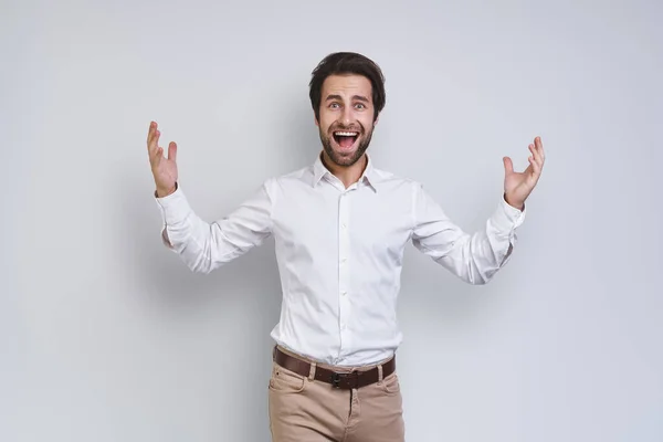 Feliz joven con camisa blanca haciendo gestos y mirando a la cámara mientras está de pie sobre un fondo gris — Foto de Stock