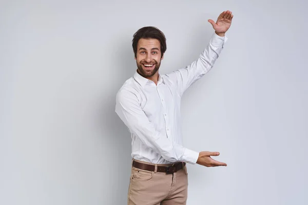 Jovem feliz em camisa branca olhando para a câmera e gesticulando enquanto está de pé contra o fundo cinza — Fotografia de Stock