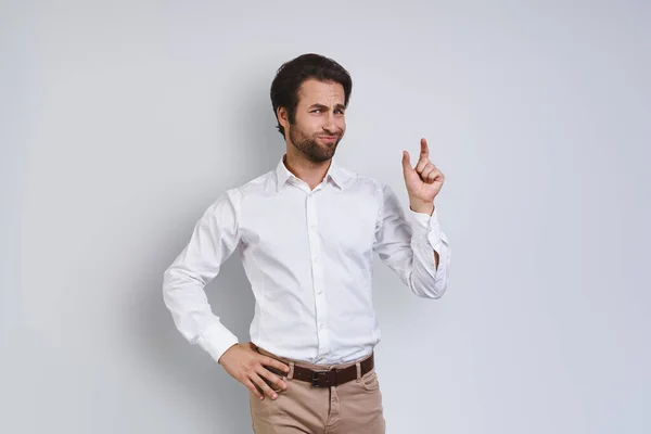 Ontevreden jongeman in wit shirt kijkend naar camera en gebaar terwijl hij tegen een grijze achtergrond staat — Stockfoto