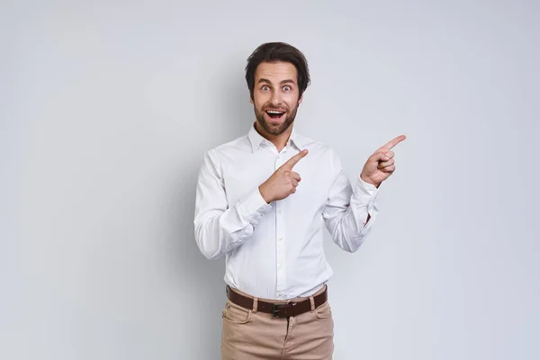 Bonito jovem sorridente homem em camisa branca olhando para a câmera e apontando para longe, enquanto em pé contra fundo cinza — Fotografia de Stock