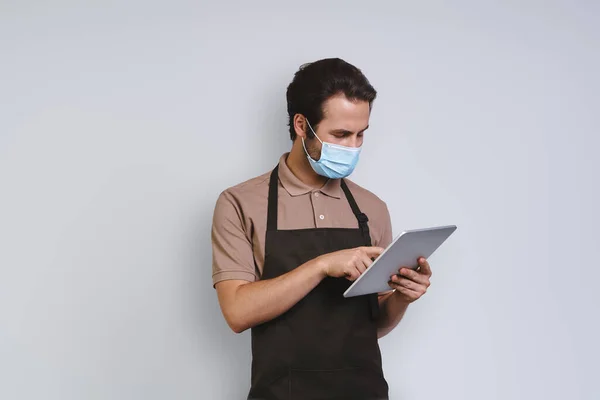 Handsome young man in apron wearing protective face and using digital tablet while standing against gray background — Stock Photo, Image