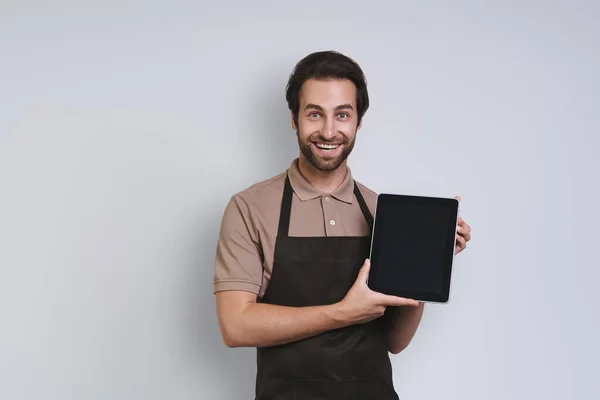 Веселый молодой человек в фартуке показывает свой цифровой планшет и улыбается стоя на сером фоне — стоковое фото