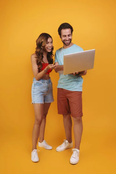 Full length of beautiful young couple using laptop and smiling while standing against yellow background — Stock Photo, Image