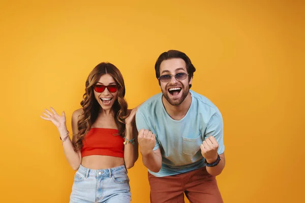 Hermosa pareja joven en gafas graduadas divirtiéndose mientras está de pie sobre el fondo amarillo —  Fotos de Stock