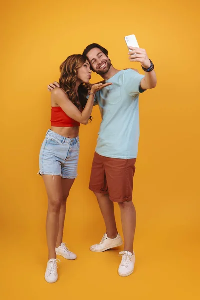 Full length of beautiful young smiling couple making selfie while standing against yellow background — Stock Photo, Image