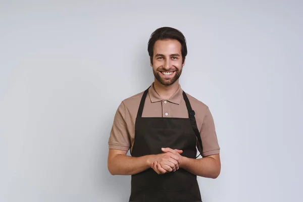 Handsome young man in apron looking at camera and keeping hands clasped while standing against gray background — Stock Photo, Image