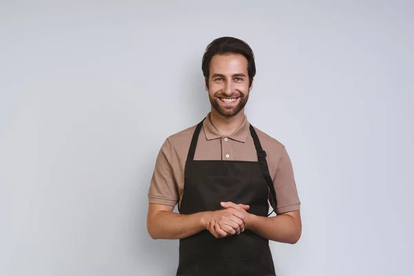 Stilig ung man i förkläde tittar på kameran och hålla händerna knäppta när du står mot grå bakgrund — Stockfoto