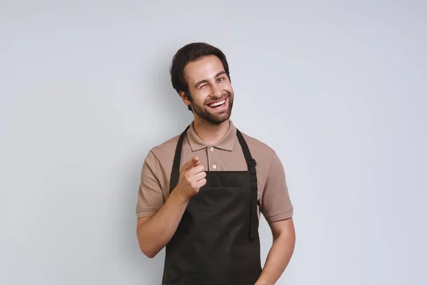 Handsome young man in apron looking at camera and pointing you while standing against gray background — Stock Photo, Image