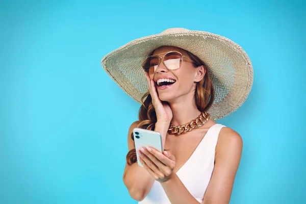 Aufgeregte junge Frau mit elegantem Hut und Handy vor blauem Hintergrund — Stockfoto