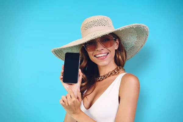 Mooie jonge vrouw in elegante hoed tonen haar mobiele telefoon terwijl staan tegen een blauwe achtergrond — Stockfoto