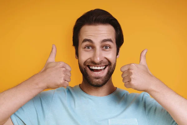 Porträtt av glad ung man i casual kläder tittar på kameran och visar tummen upp när du står mot gul bakgrund — Stockfoto