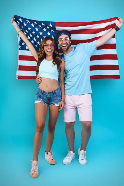 Amerikan bayrağı taşıyan ve mavi arka planda dururken gülümseyen güzel genç bir çift. — Stok fotoğraf